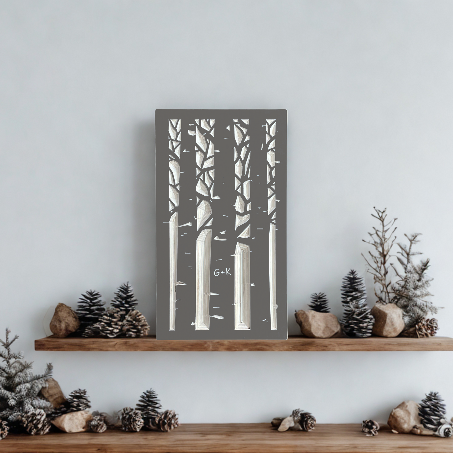 Decorative wooden wall art with taupe gray silhouettes of an aspen forest with a couple's initials carved in a trunk. Shown on a wooden shelf with accessories against white wall.