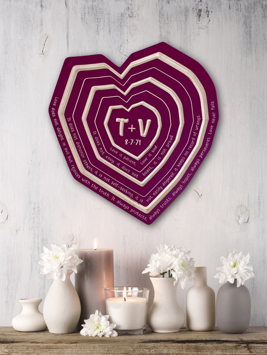 Heart-shaped decorative wall art with text on a wooden shelf with candles and flowers.
