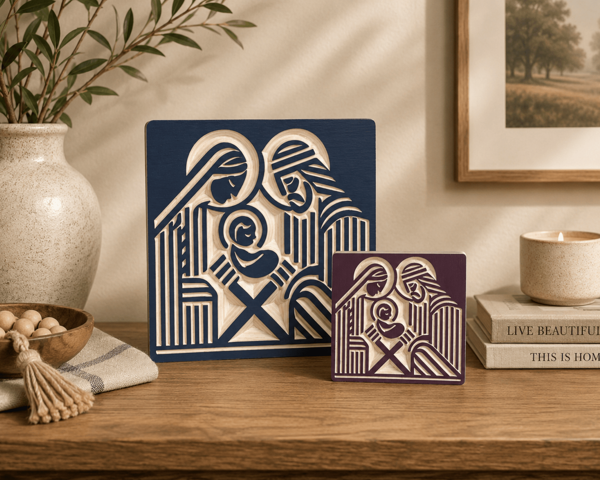 Two decorative wall art panels with a stylized Holy Family design, displayed on a wooden shelf in a styled home interior. One panel is dark navy blue and the other is deep burgundy, both with cream cutout artwork.