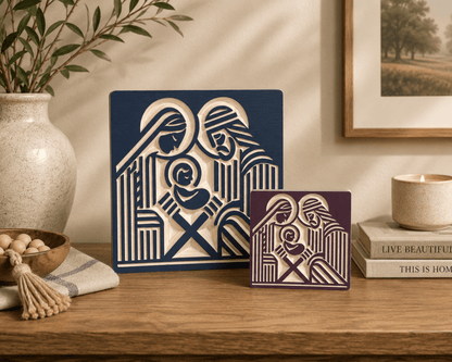 Two decorative wall art panels with a stylized Holy Family design, displayed on a wooden shelf in a styled home interior. One panel is dark navy blue and the other is deep burgundy, both with cream cutout artwork.