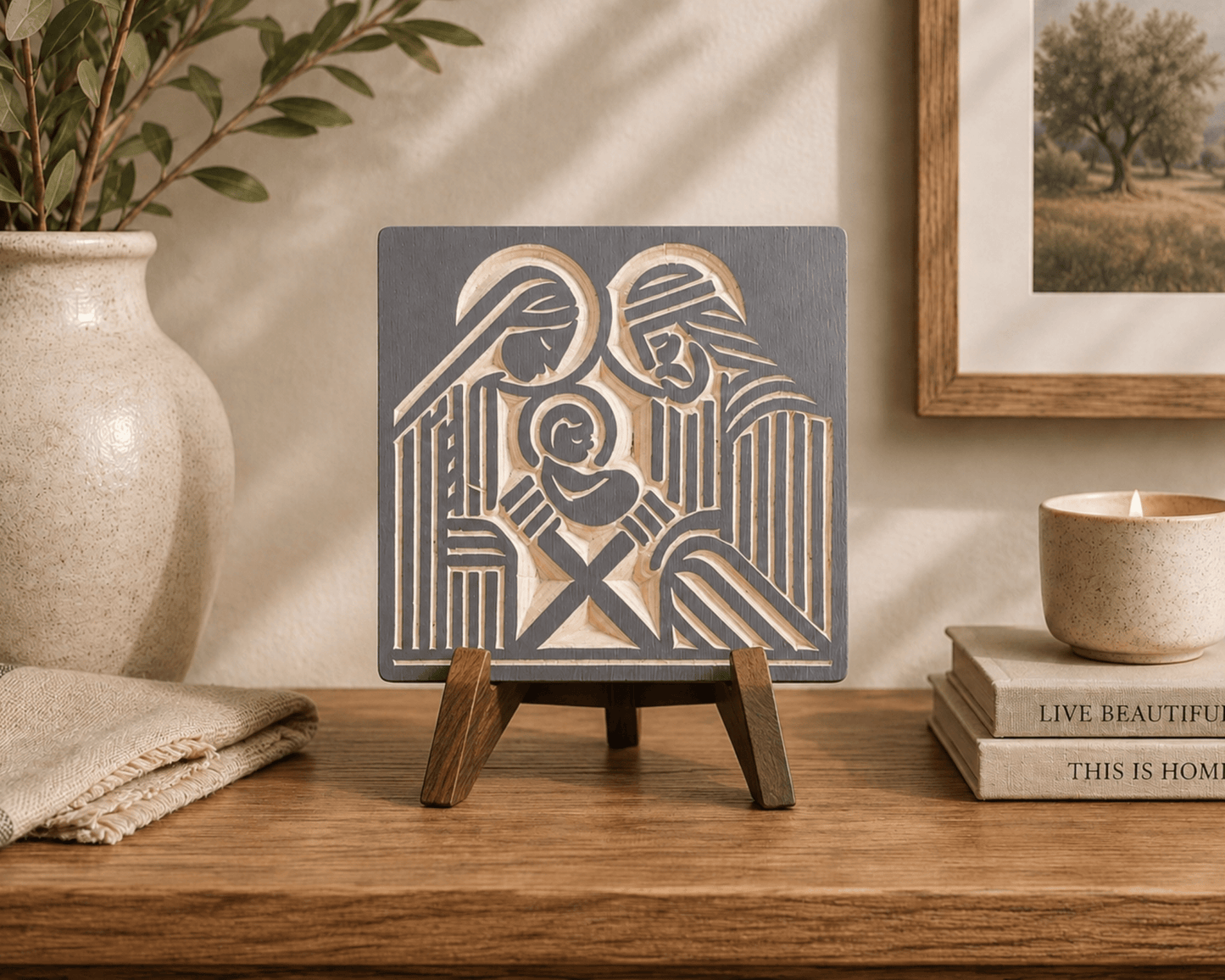 A square decorative wall art plaque featuring a stylized nativity scene in dark charcoal gray and warm beige, displayed on a small wooden easel stand on a wooden table. The scene is styled in a warm, rustic room with a beige ceramic vase, a candle, and books in the background.