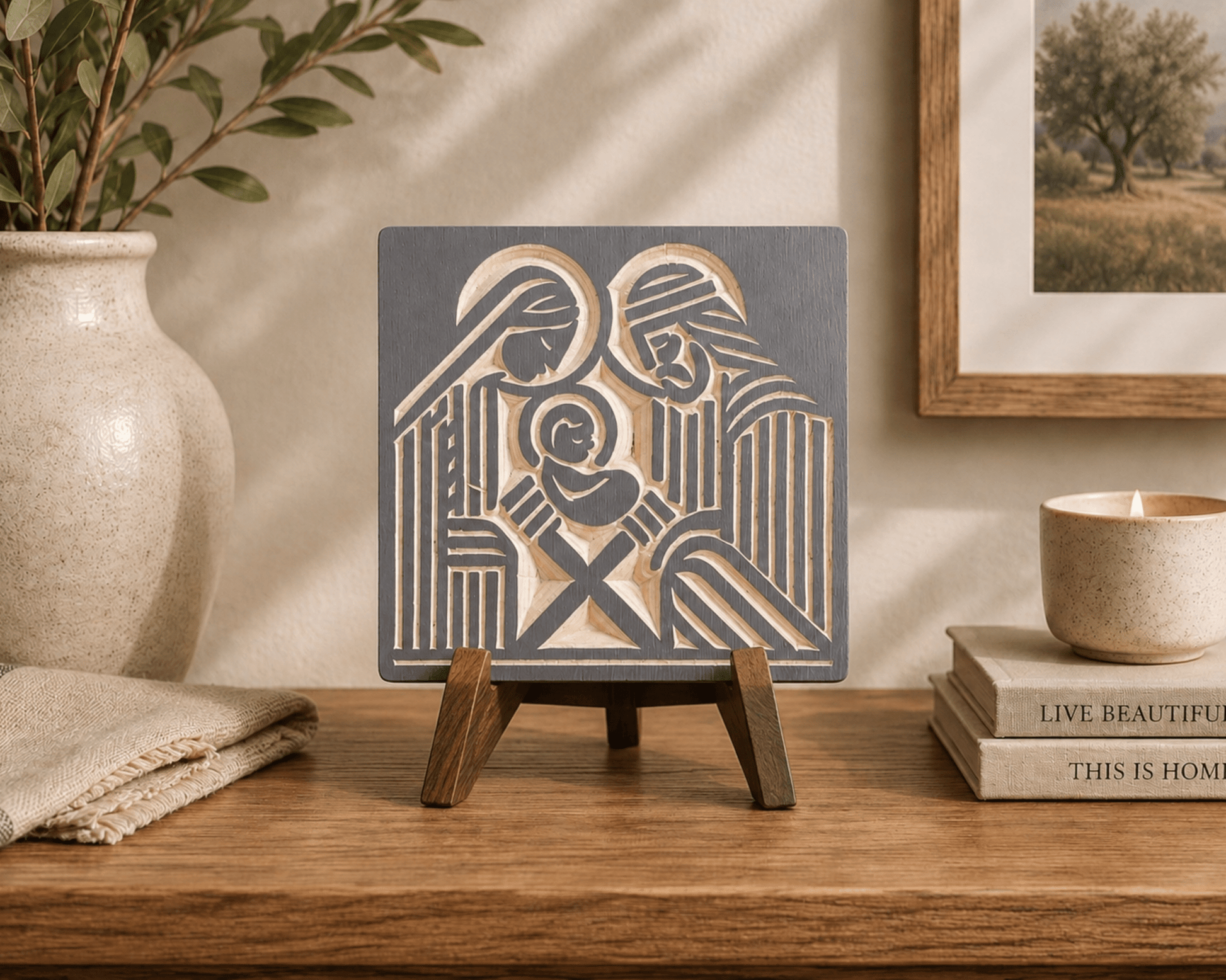 A square decorative wall art plaque featuring a stylized nativity scene in dark charcoal gray and warm beige, displayed on a small wooden easel stand on a wooden table. The scene is styled in a warm, rustic room with a beige ceramic vase, a candle, and books in the background.