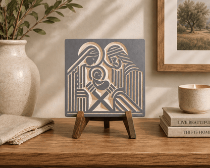 A square decorative wall art plaque featuring a stylized nativity scene in dark charcoal gray and warm beige, displayed on a small wooden easel stand on a wooden table. The scene is styled in a warm, rustic room with a beige ceramic vase, a candle, and books in the background.