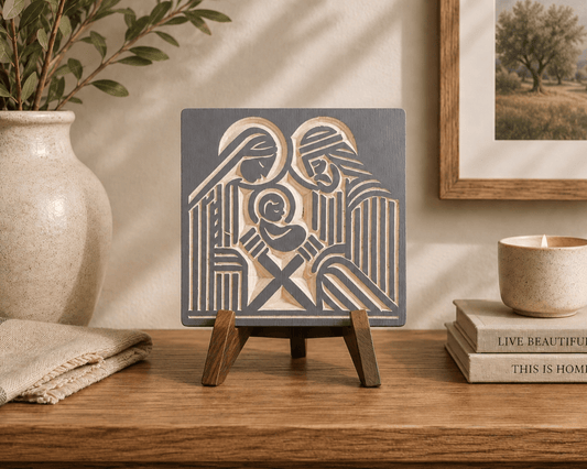 A square decorative wall art plaque featuring a stylized nativity scene in dark charcoal gray and warm beige, displayed on a small wooden easel stand on a wooden table. The scene is styled in a warm, rustic room with a beige ceramic vase, a candle, and books in the background.