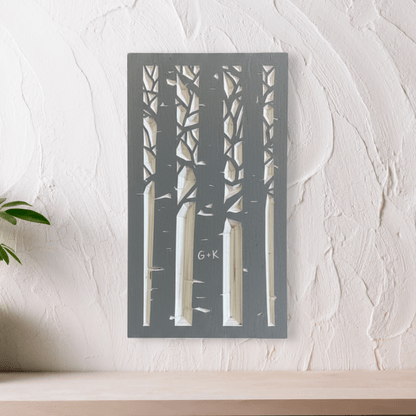 Wooden wall art with driftwood gray silhouettes of an aspen forest with a couple's initials carved in a trunk. Shown on above a wooden shelf with  against white wall. 