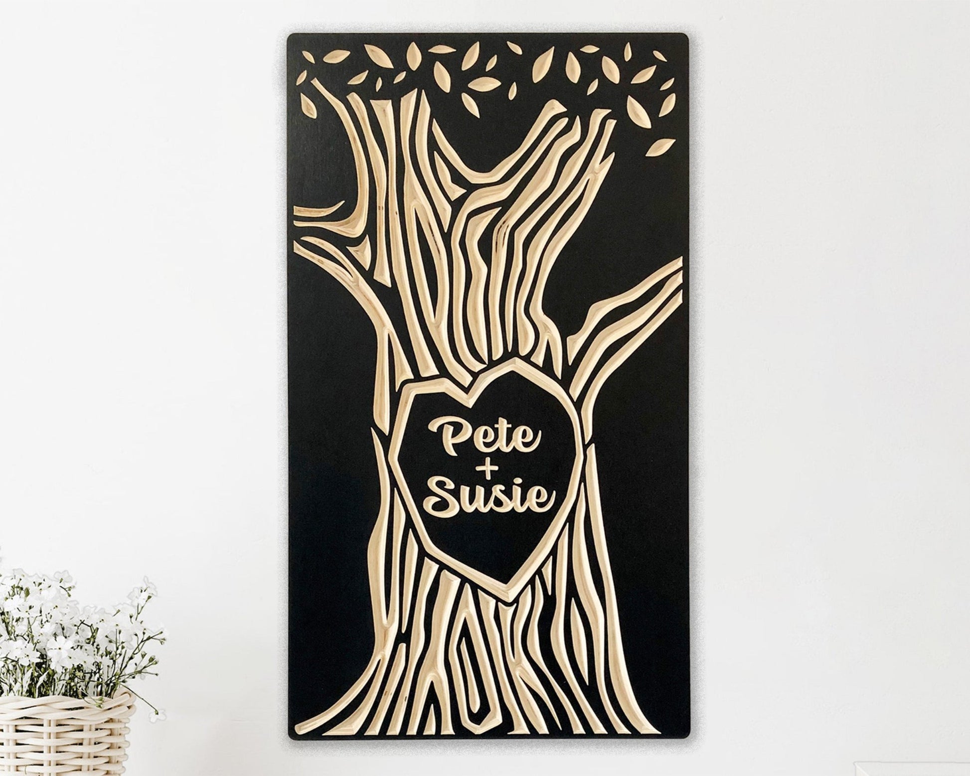 Solid wood panel, painted black. Tree trunk with a heart shape in the center and canopy of leaves is carved as the design. Personalized with two names, like a couple carving their names on a tree.