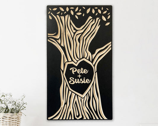 Solid wood panel, painted black. Tree trunk with a heart shape in the center and canopy of leaves is carved as the design. Personalized with two names, like a couple carving their names on a tree.