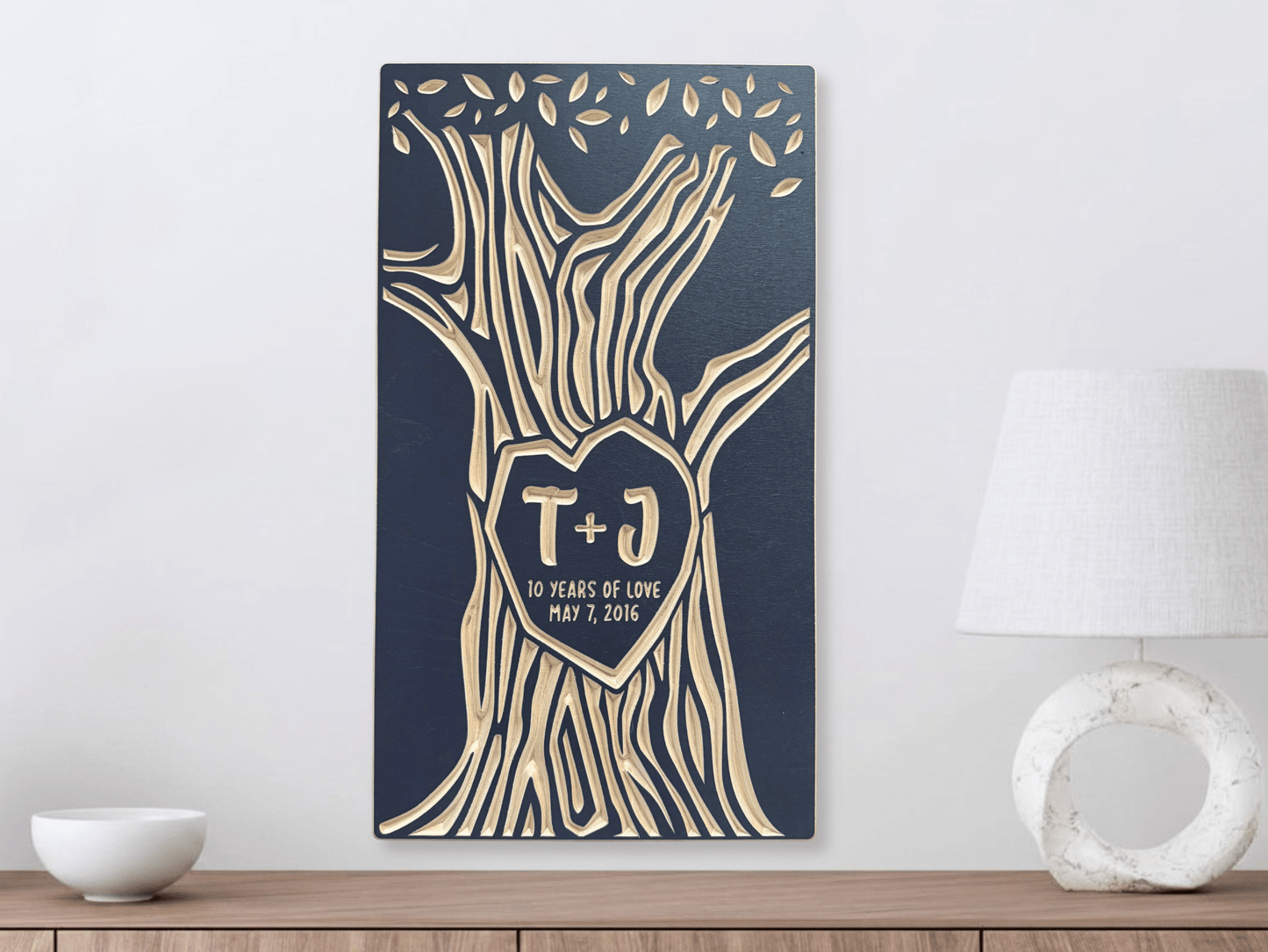 Decorative wall art with tree design and initials on a blue background, placed on a wooden surface.