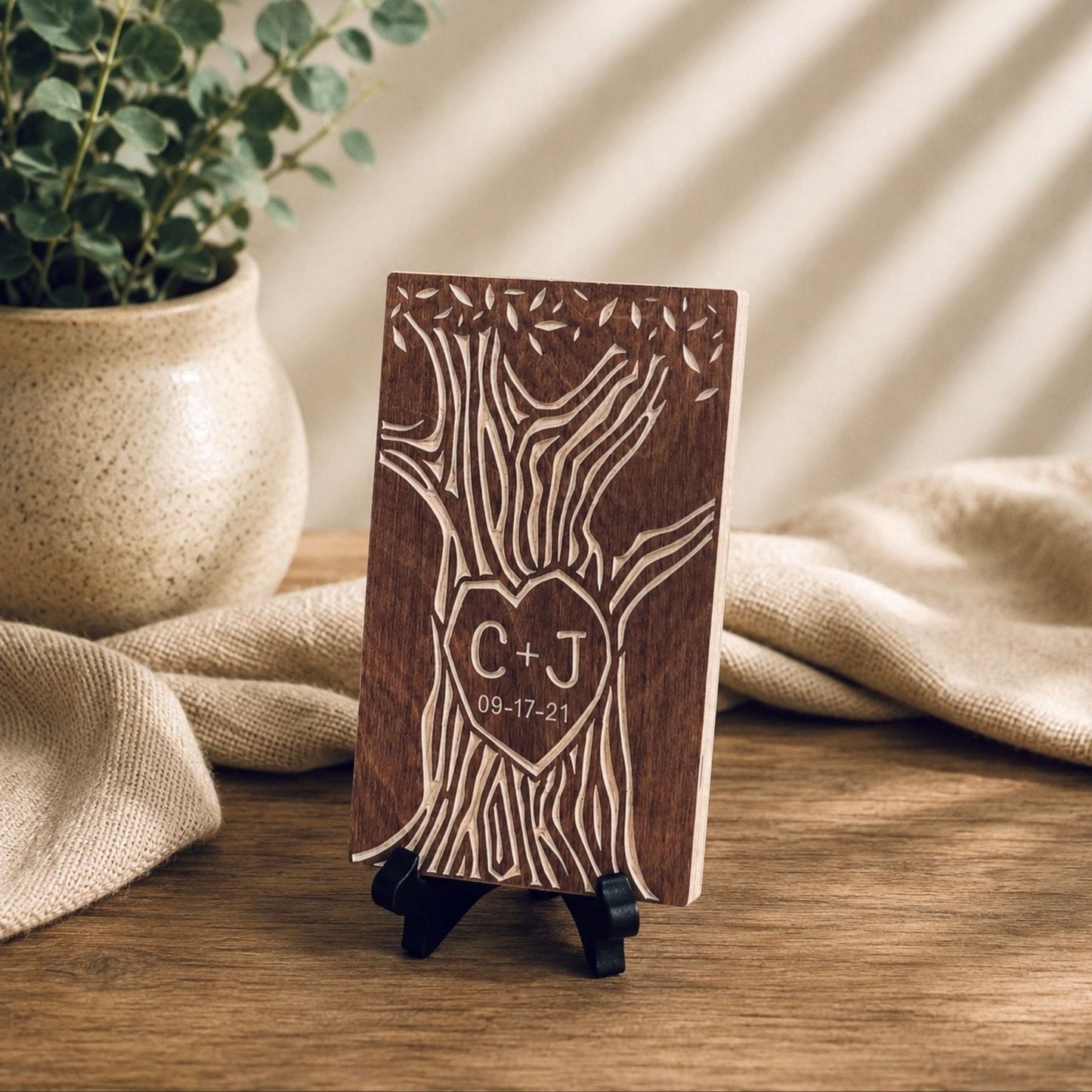 A personalized wooden plaque with an engraved tree design and a heart-shaped monogram 'C+J' with the date '09-17-21', displayed on a small black stand on a wooden table with a potted plant and beige fabric in the background.