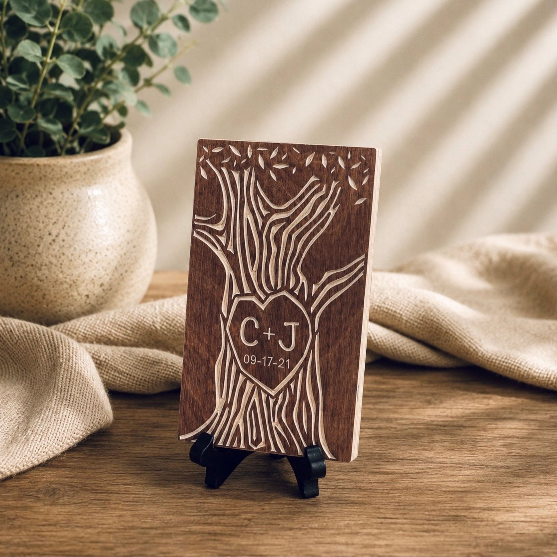 A personalized wooden plaque with an engraved tree design and a heart-shaped monogram 'C+J' with the date '09-17-21', displayed on a small black stand on a wooden table with a potted plant and beige fabric in the background.