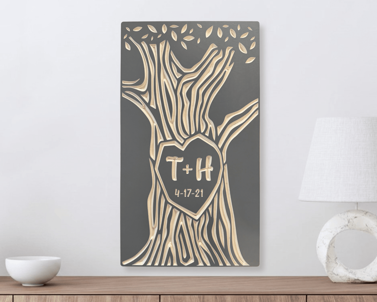 Decorative wall art with tree design and custom initials with anniversary date in taupe color option. Shown hanging on white wall above a wooden shelf.