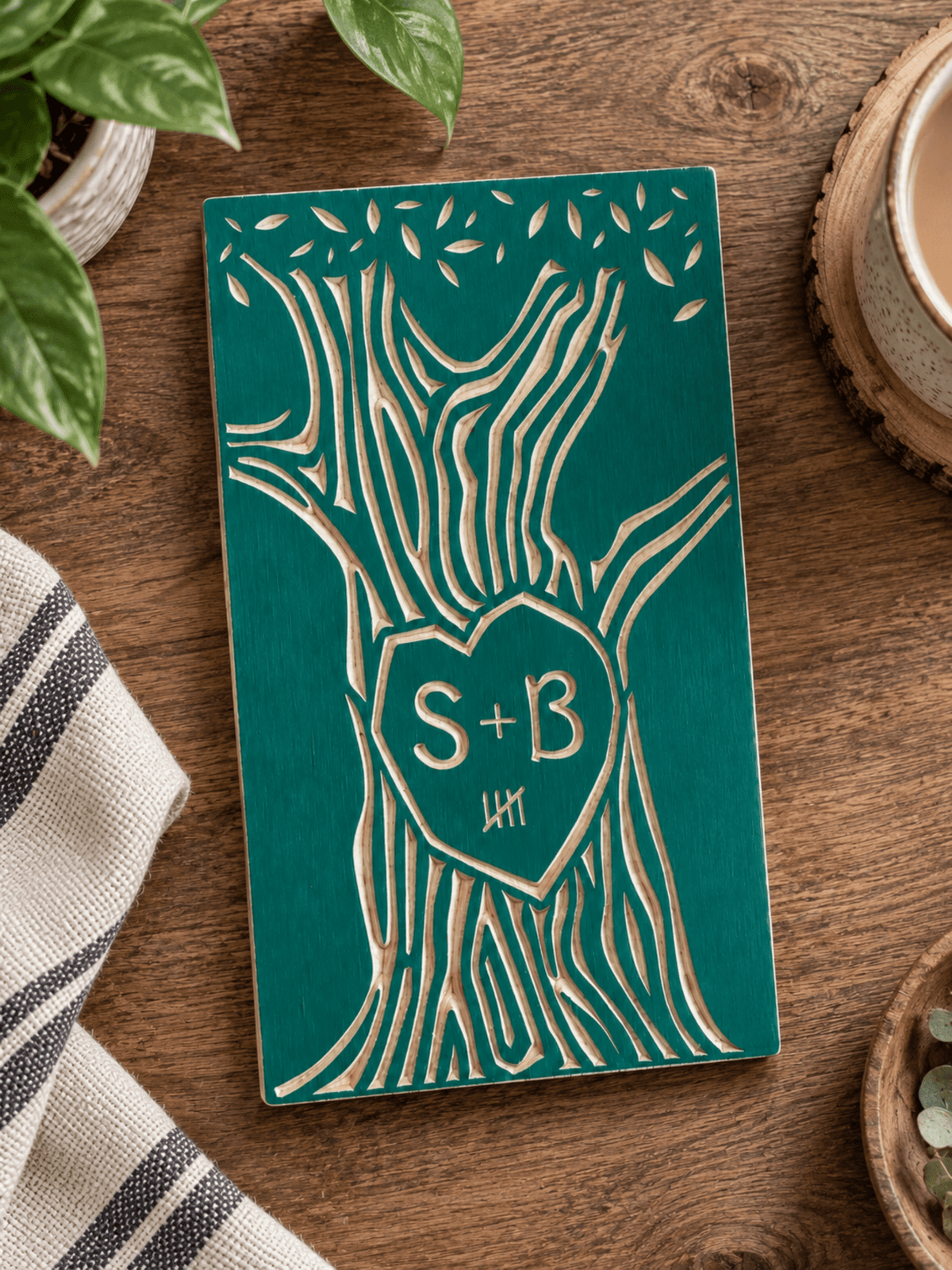 A personalized wooden plaque featuring a carved tree design with a heart-shaped center containing the initials 'S+B' and 5 tally marks, displayed on a rustic wooden tabletop with a striped cloth, plant leaves, and a ceramic cup nearby.